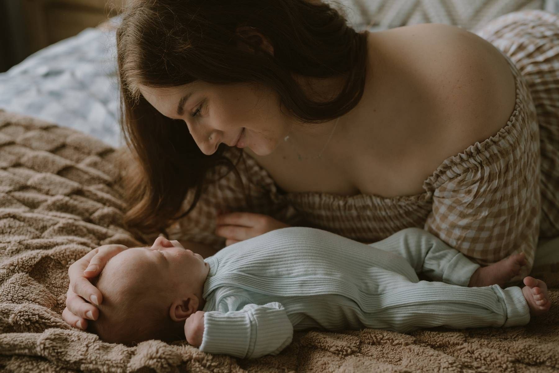 A mother tenderly gazes at her newborn baby lying on a cozy brown textured blanket on a bed.