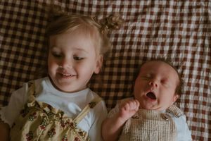 Toddler girl smiling next to yawning newborn baby lying on a brown gingham blanket.