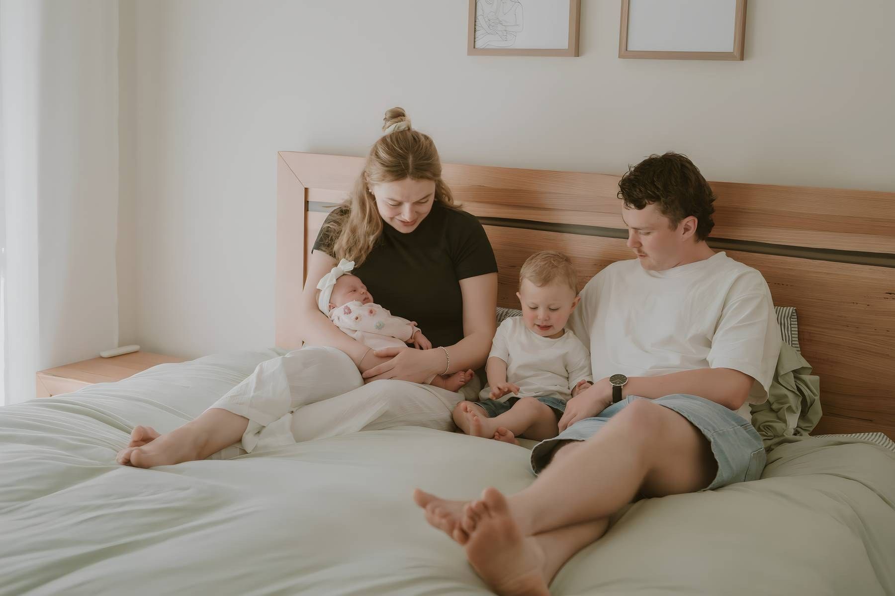Family of four sitting on a white bed, mother holding newborn baby, toddler and father beside her in a bright room.