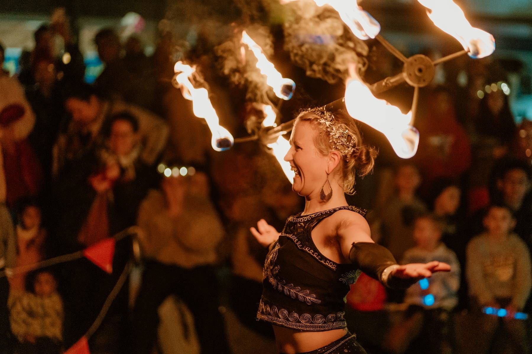 A fire dancer performs with flaming props before a captivated crowd at an outdoor night event.