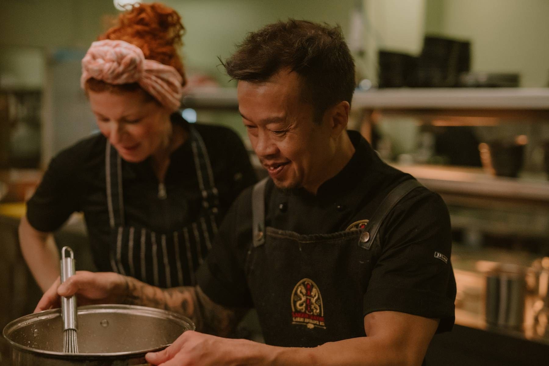 Two chefs in black aprons cooking together in a professional kitchen, smiling while preparing food.