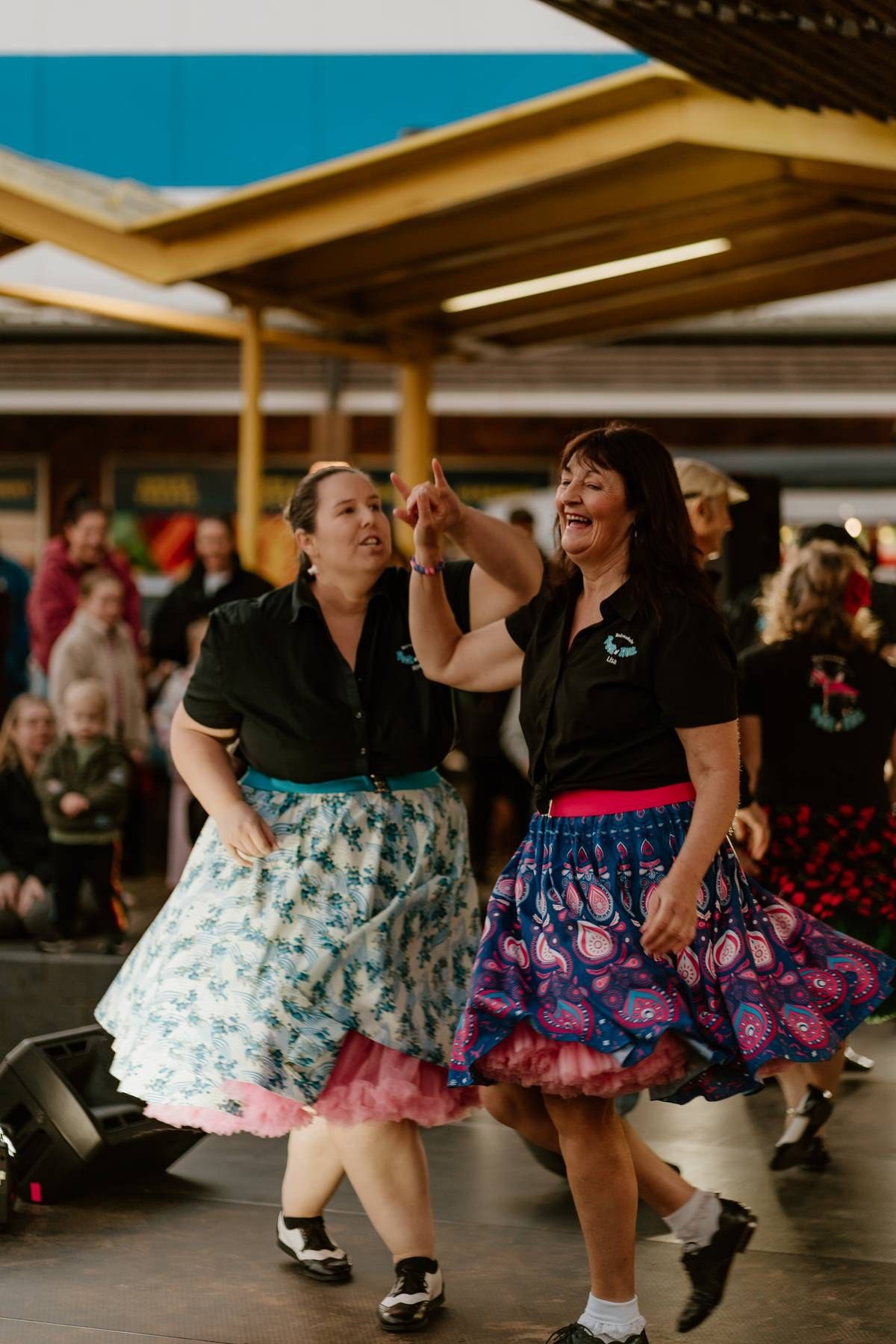 Two women in black tops and colorful swing skirts dance joyfully at a retro-themed event with an audience watching.