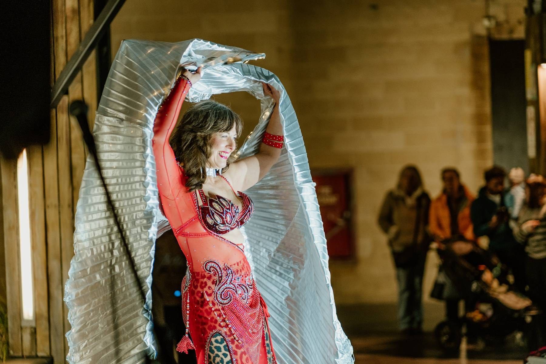 A belly dancer in red costume performs with shimmering silver wings for an audience indoors.