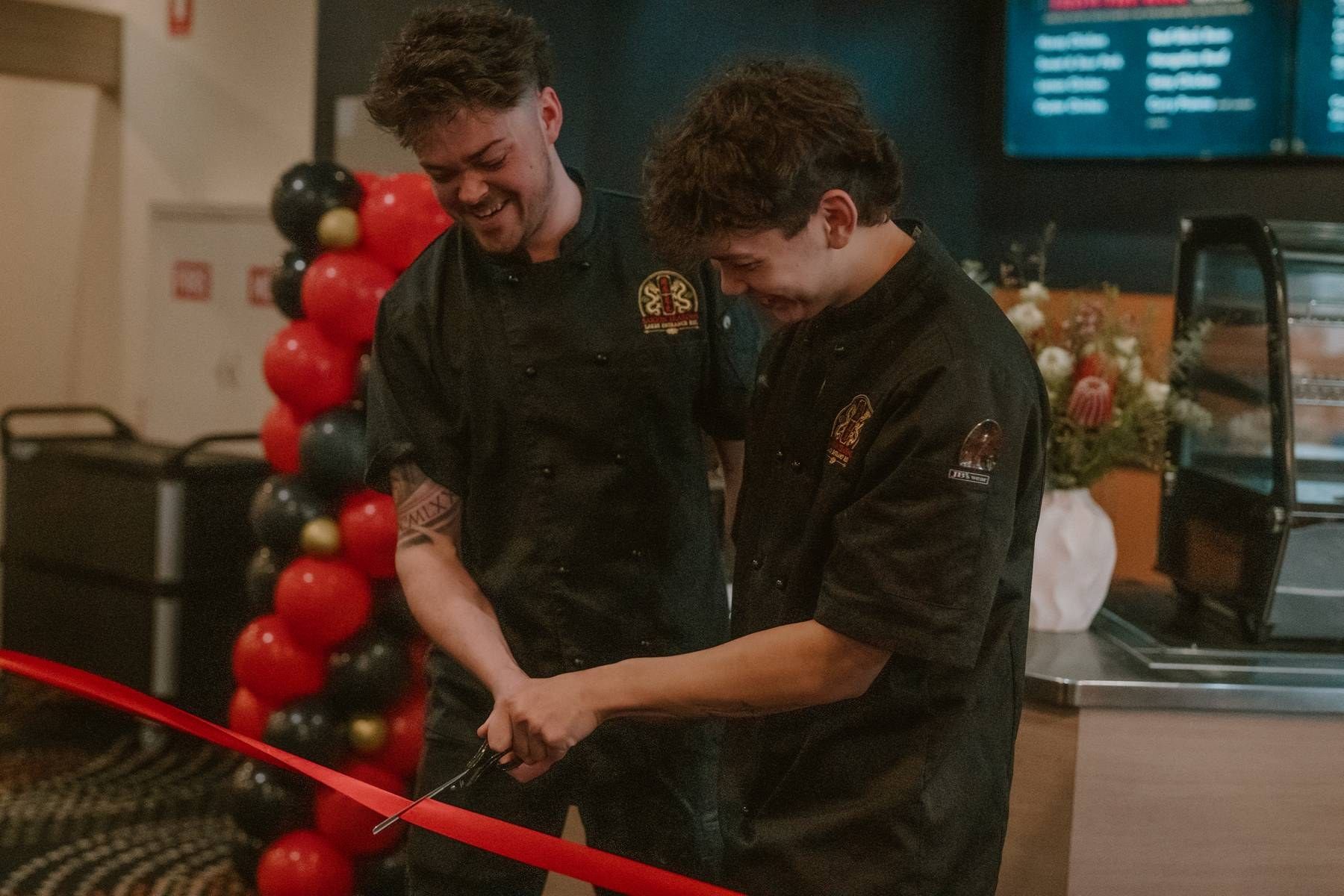 Two chefs in black uniforms cut a red ribbon at a restaurant grand opening with balloon decorations.