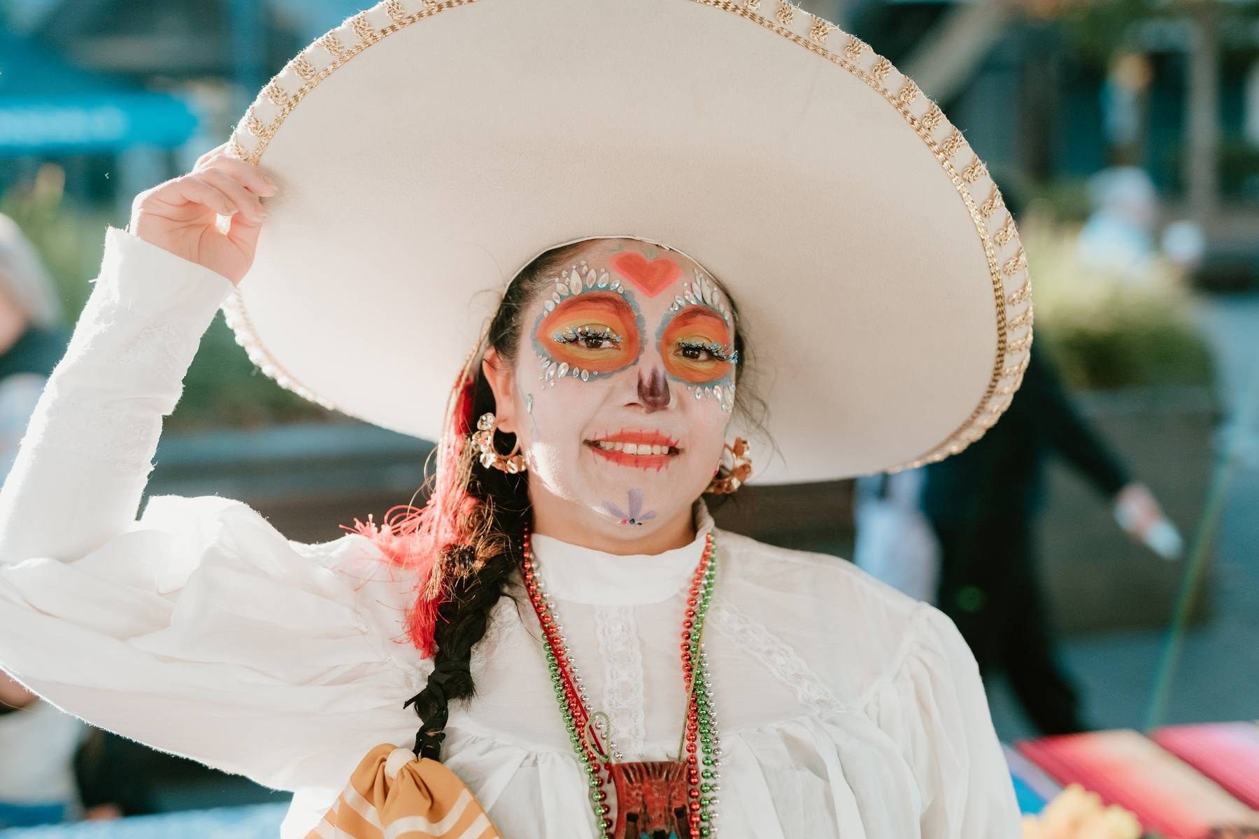 Girl in white dress and sombrero with colorful Day of the Dead face paint smiles at camera.