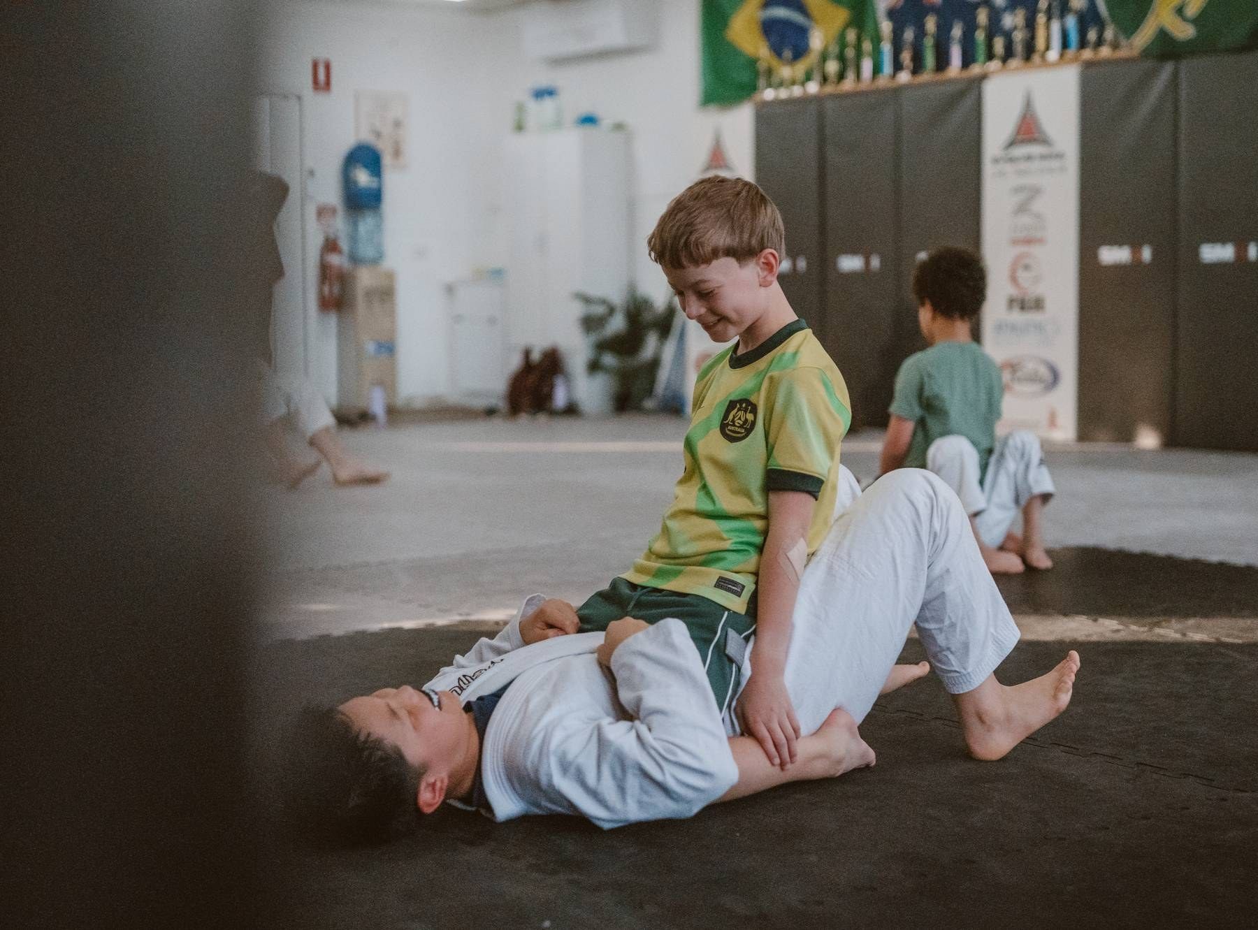 Children practicing Brazilian Jiu-Jitsu grappling techniques on a mat in a martial arts gym.