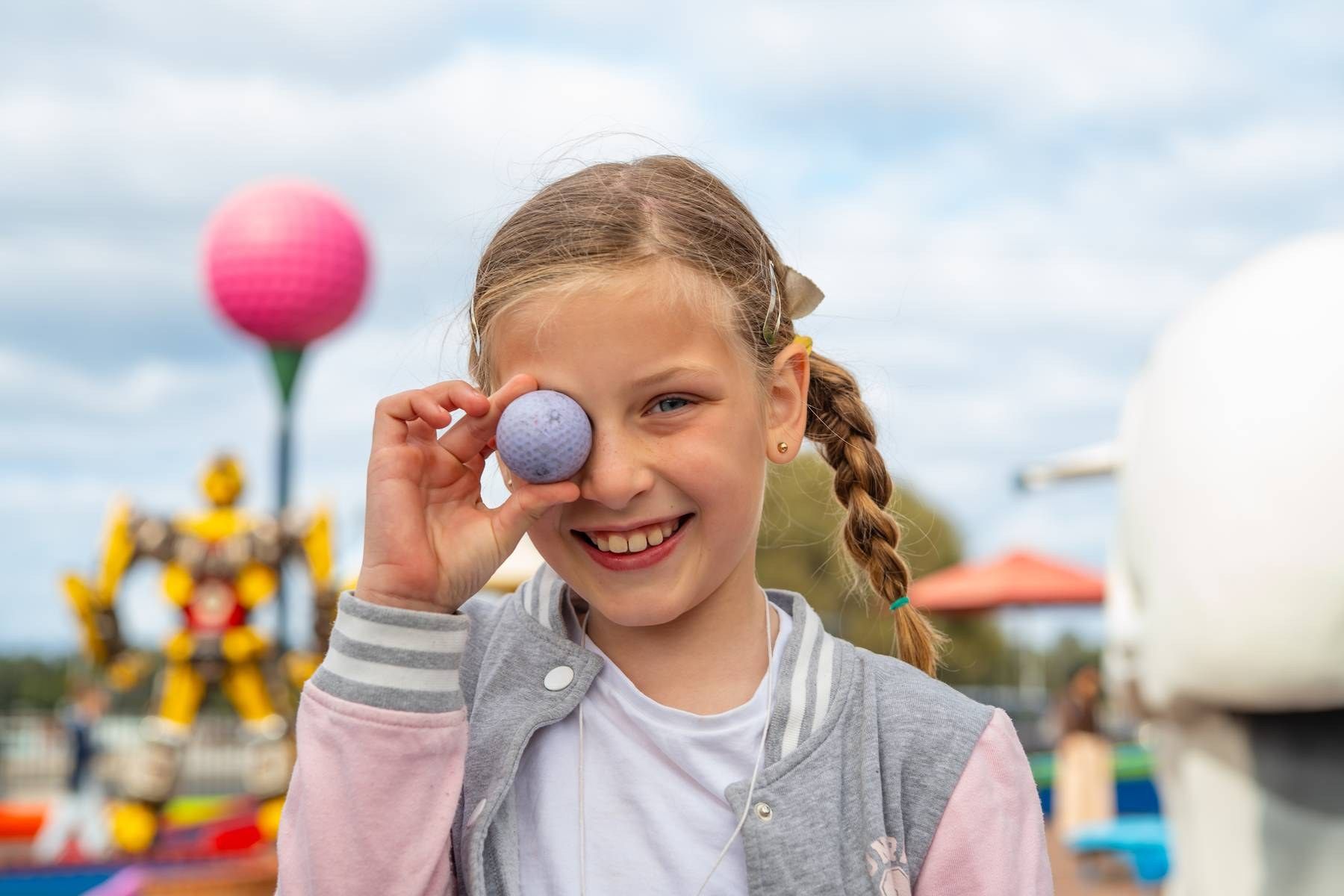 Happy girl holding purple ball over her eye at colorful amusement park on cloudy day.