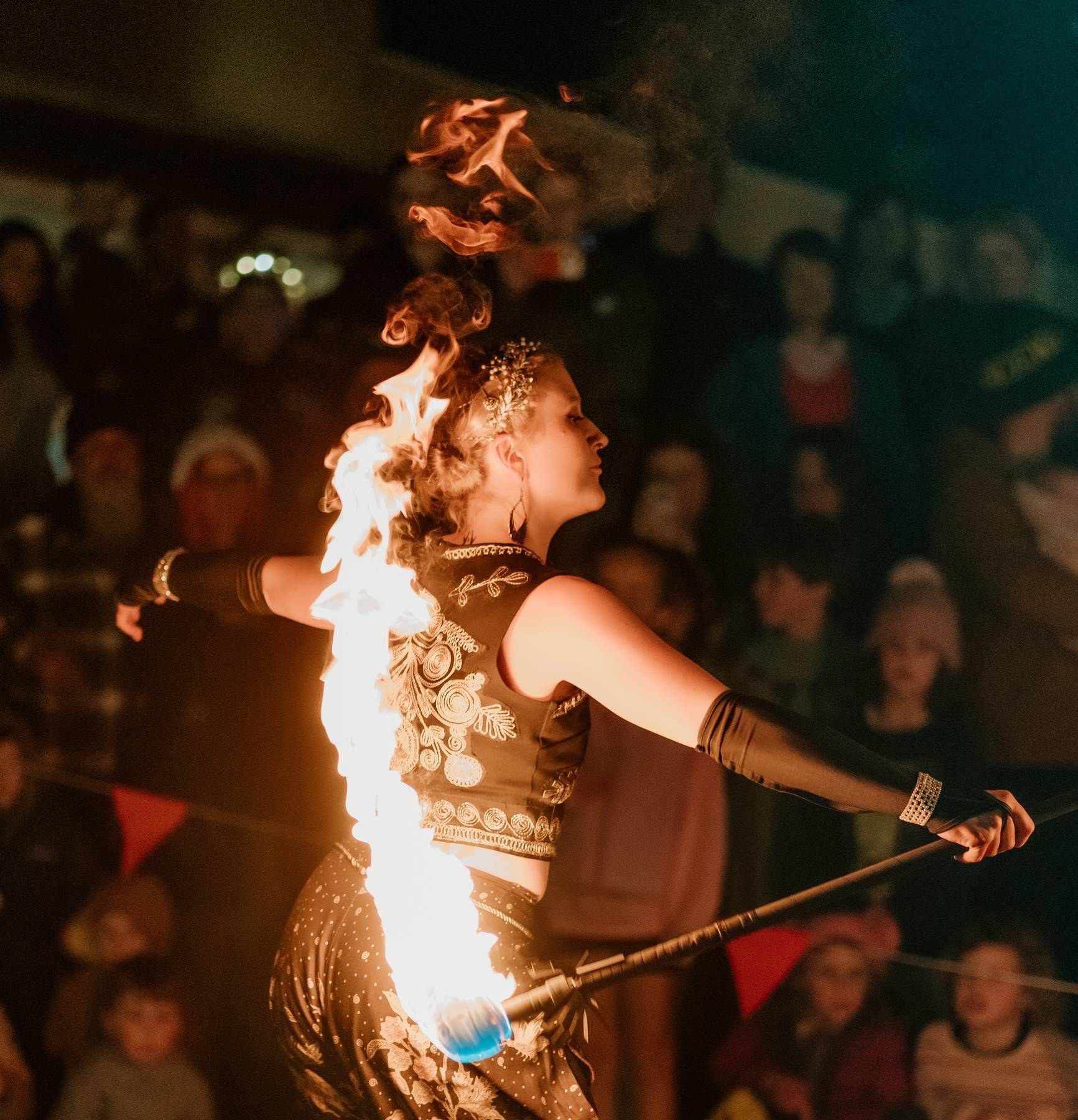 A fire dancer performs with flaming batons before a crowd at night, wearing ornate costume with flames illuminating the scene.
