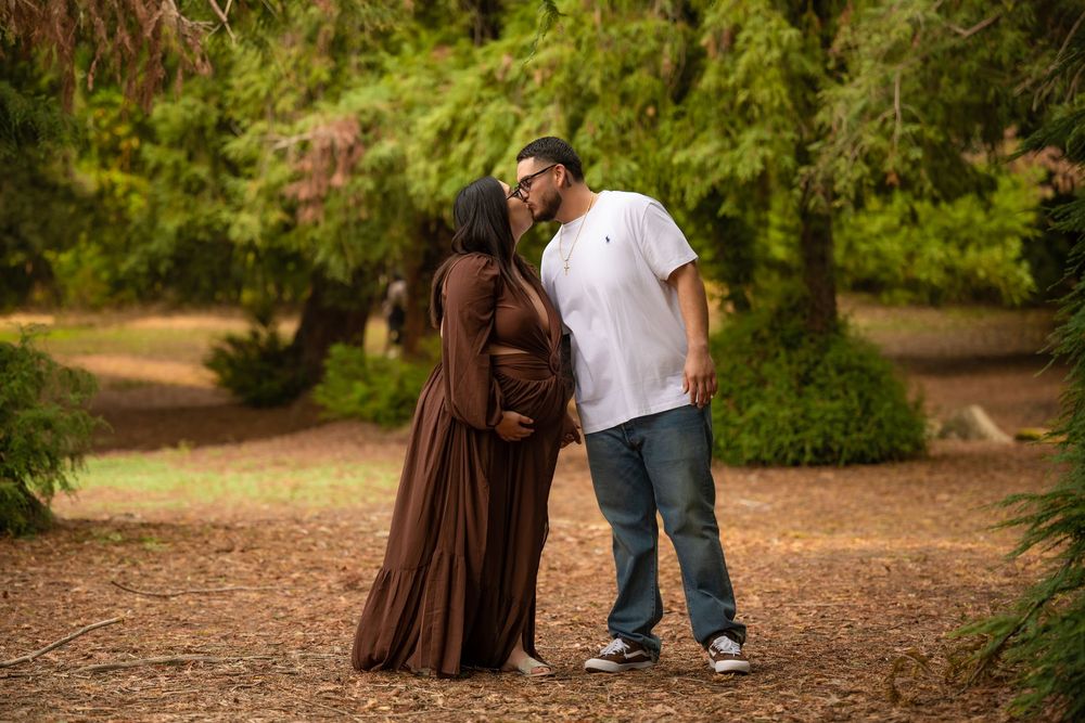Expectant couple sharing a kiss outdoors beneath green trees during a maternity photography session.