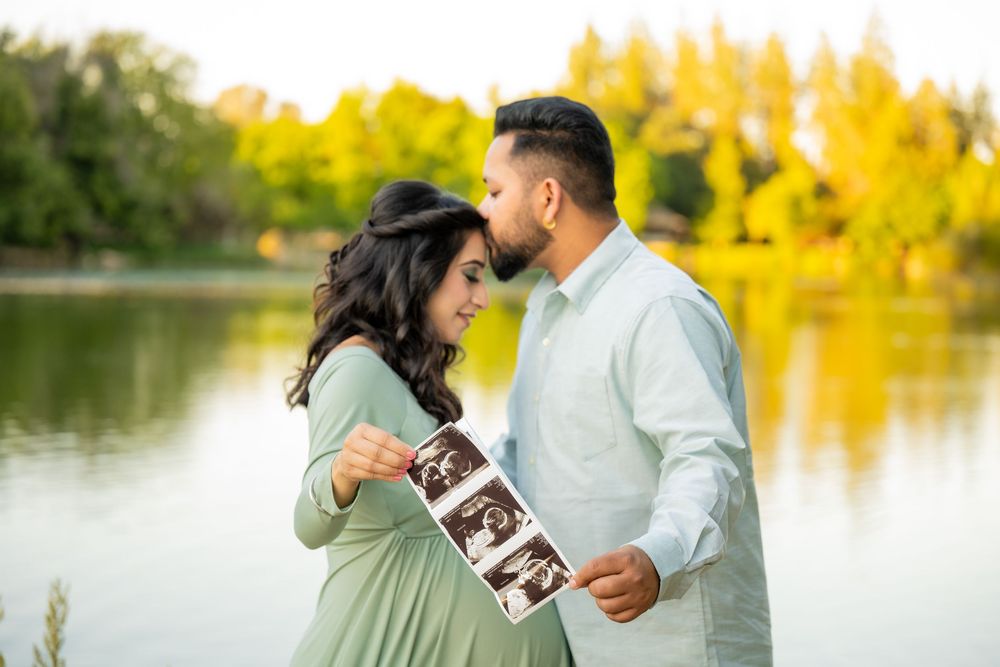 Expectant couple sharing a tender moment by a lake while holding ultrasound photographs of their baby.