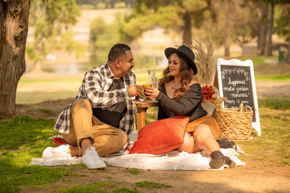 Couple enjoying a romantic outdoor picnic with pillows and flowers in a sunlit park.
