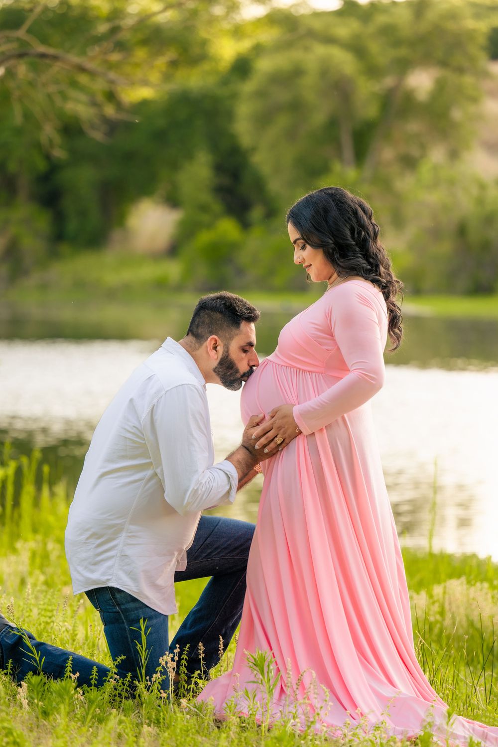 Expectant father kneeling to kiss his partner's baby bump as she wears a flowing pink maternity gown beside a lake.