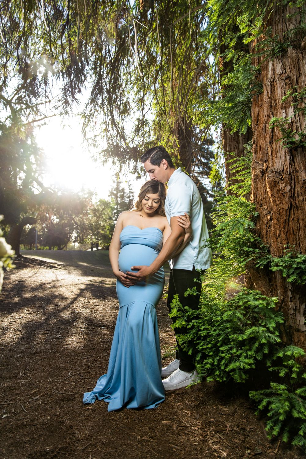 Expectant couple posing beneath tall trees, with the mother in a flowing blue maternity gown.