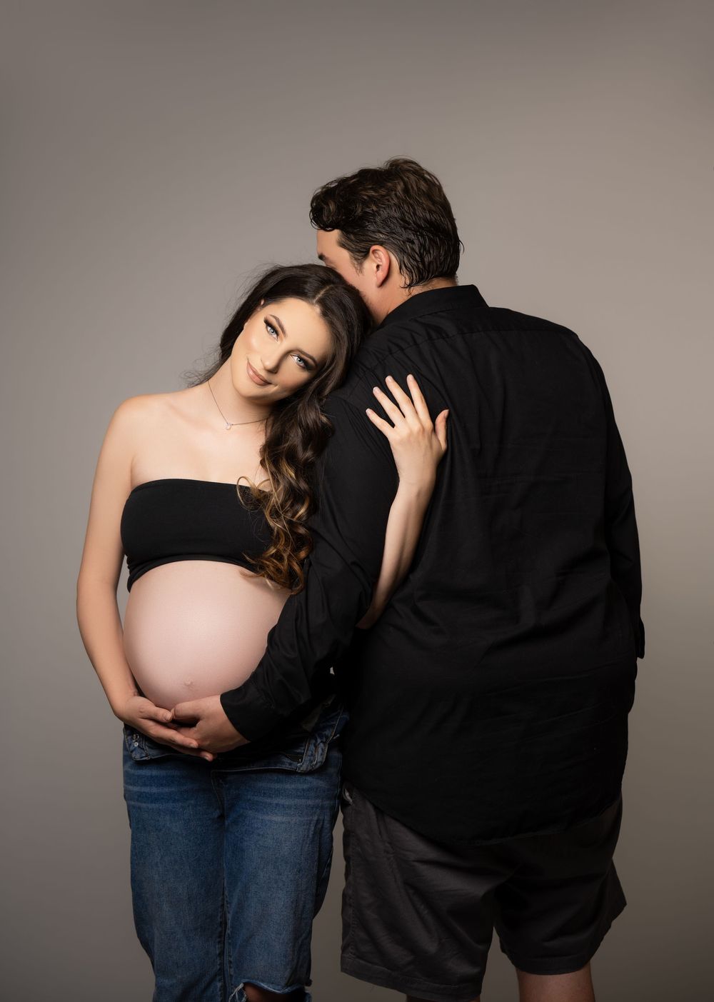 Expectant couple embracing during a studio maternity photoshoot, with the mother gently holding her baby bump.