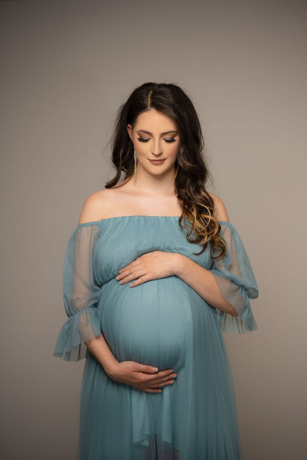 Pregnant woman in a flowing teal blue gown gently cradling her baby bump during a studio maternity portrait session.