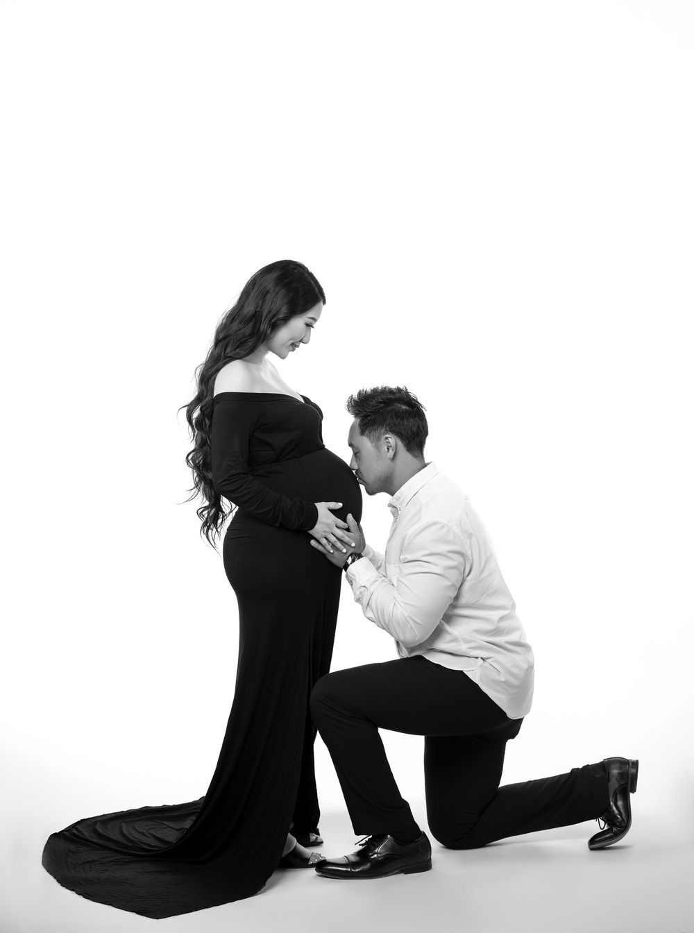 Black-and-white maternity portrait of an expectant mother in a long black dress as her partner kneels to kiss her baby bump.
