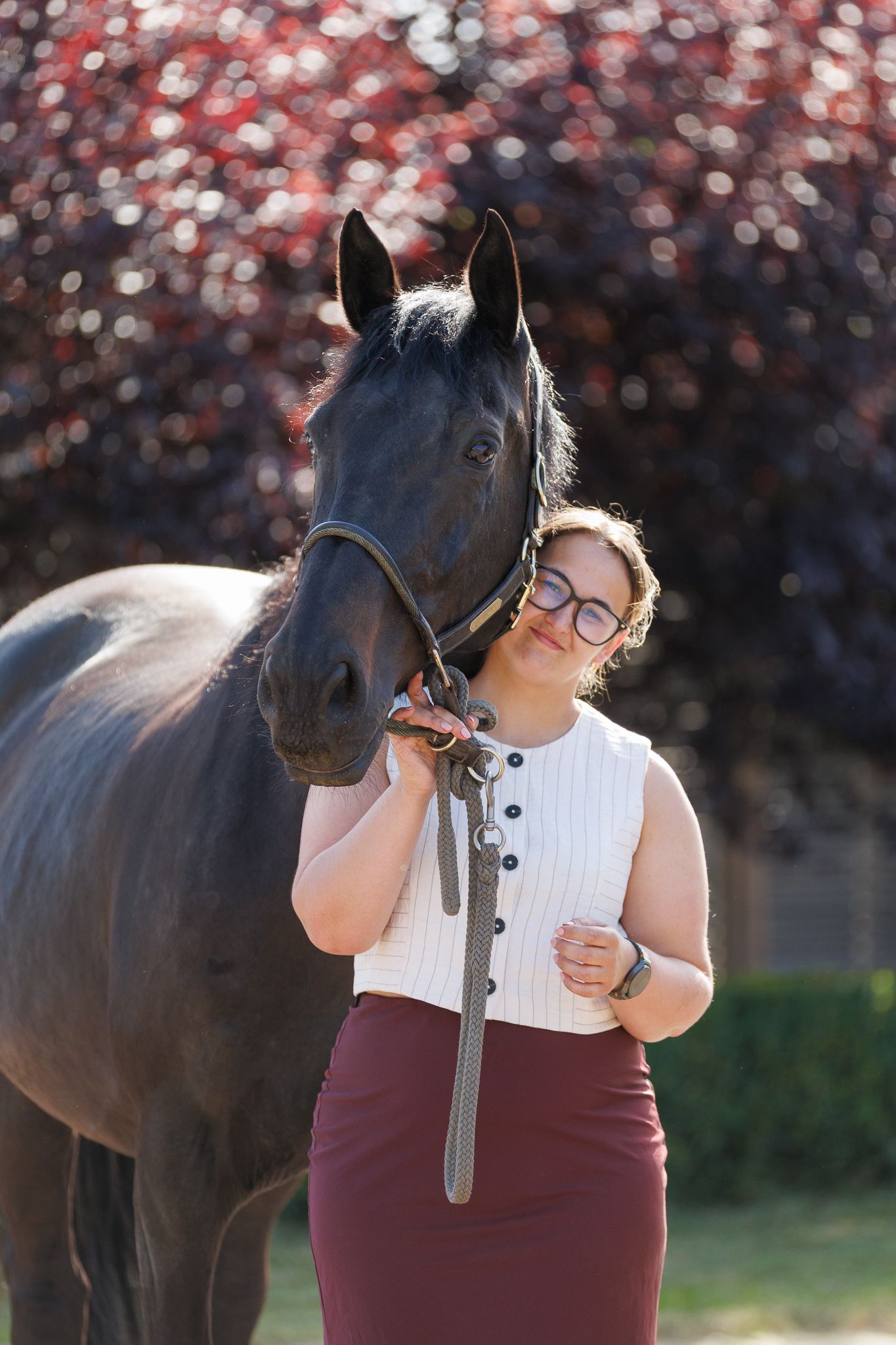 Woman in glasses, white sleeveless top and burgundy skirt rests her head against a dark horse in a bridle outdoors near red-leaved trees.