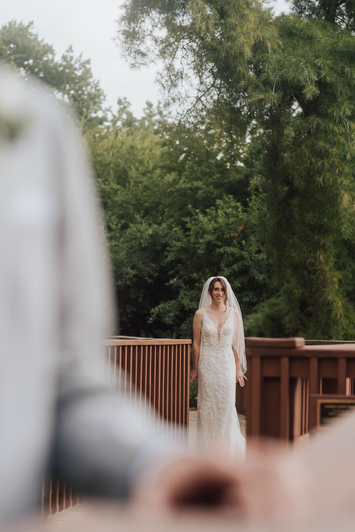 Bride in lace wedding dress and veil stands on wooden deck surrounded by lush green trees during outdoor wedding ceremony.