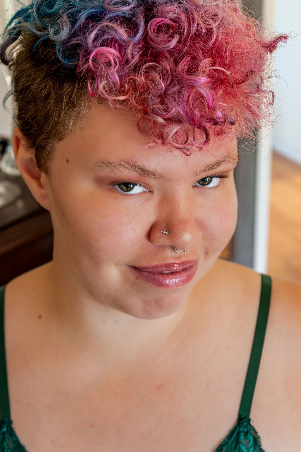 Close-up portrait of a young woman with vibrant pink and blue curly hair and a nose piercing smiling softly.