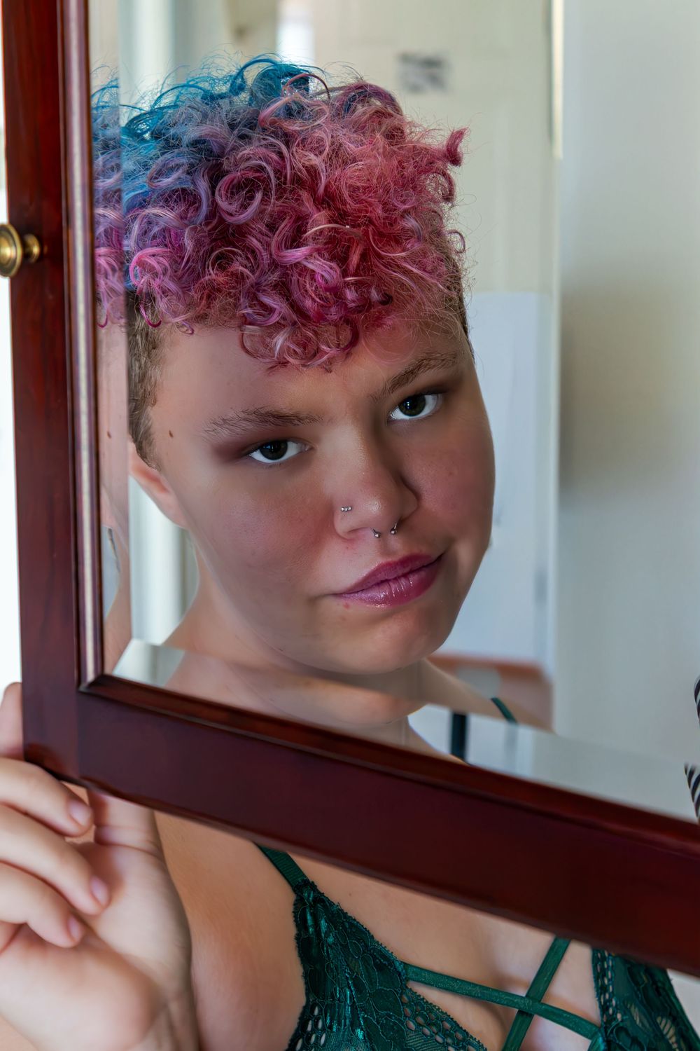 Young woman with colorful pink and blue curly hair poses beside a wooden framed mirror indoors.