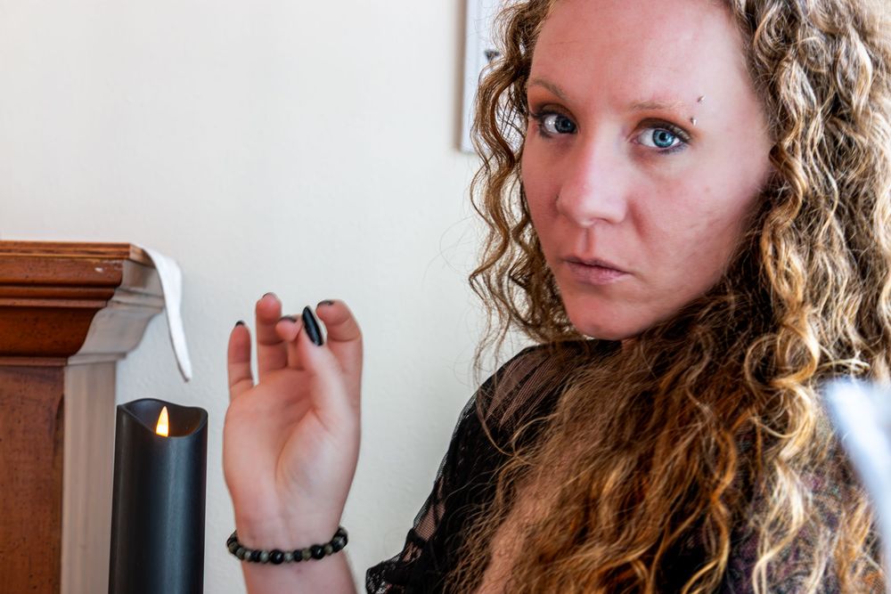 Curly-haired woman with blue eyes and facial piercings holding dark nail polish near candlestick.
