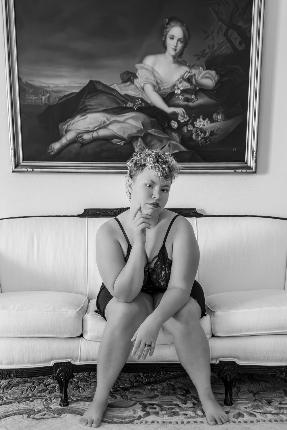 Woman in black lingerie sitting on white sofa beneath classical oil painting in black and white photo.