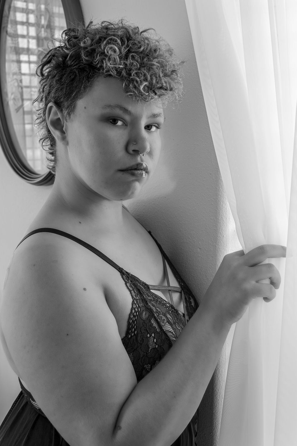 Black and white portrait of a young woman with curly short hair holding a sheer curtain near a window.