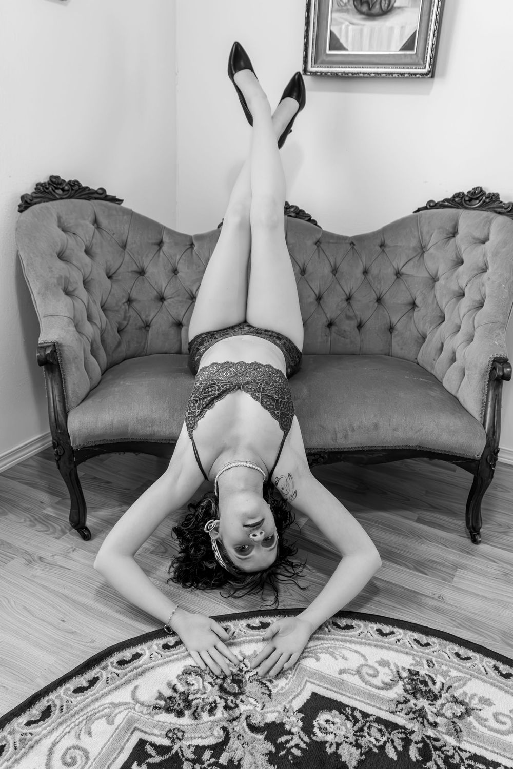 Black and white photo of a woman in lingerie lying on a rug with legs raised against a vintage tufted sofa.