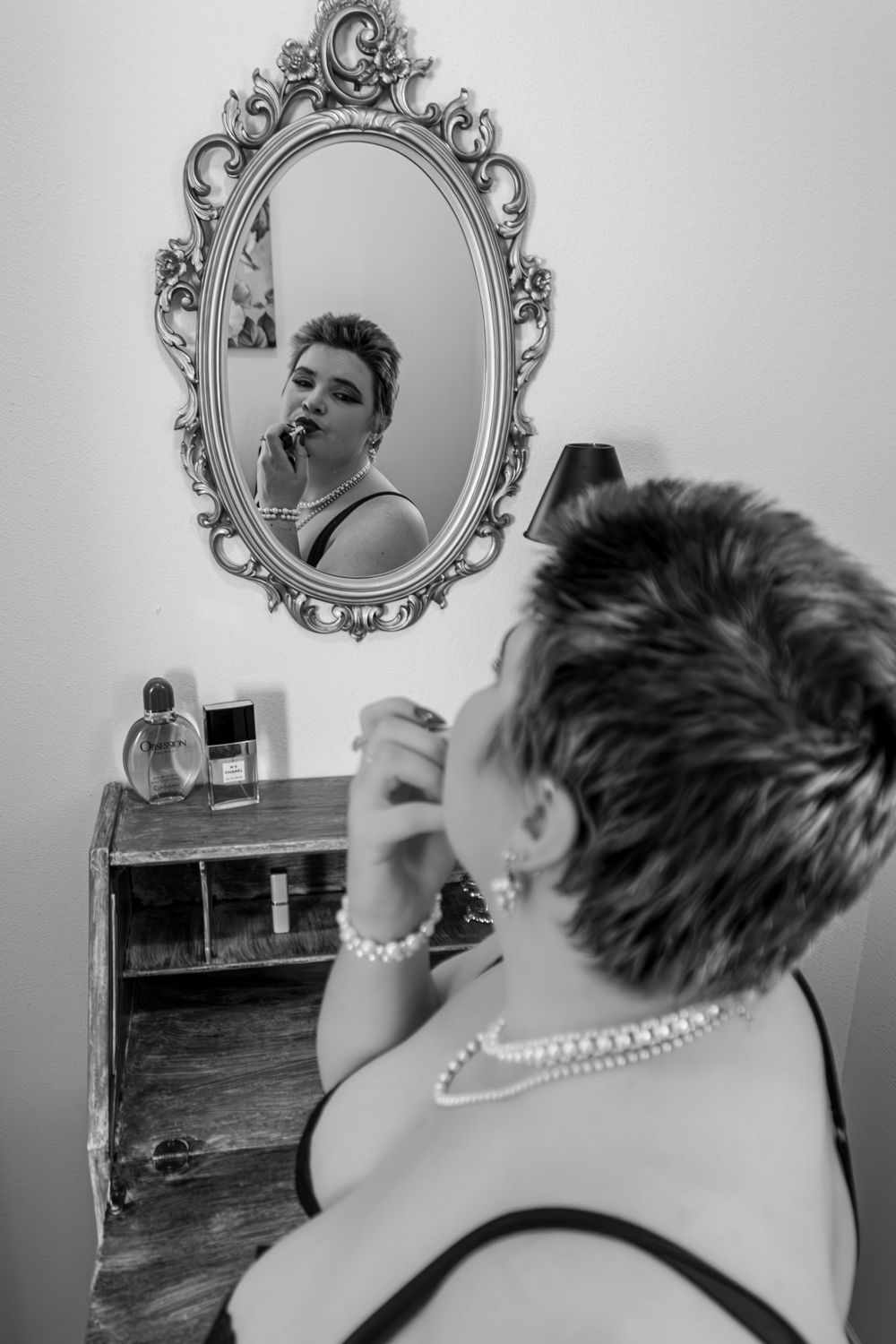 Woman applying lipstick reflected in ornate vintage mirror with perfume bottles on wooden shelf.