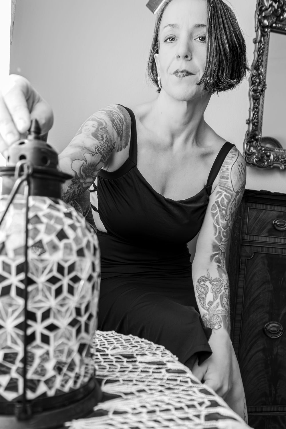 Black and white portrait of tattooed woman in black dress reaching toward decorative lantern.
