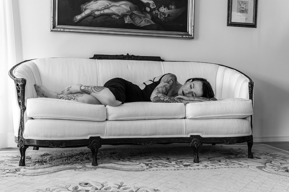 Black and white photo of a tattooed woman resting on an ornate white vintage sofa beneath a classical painting.