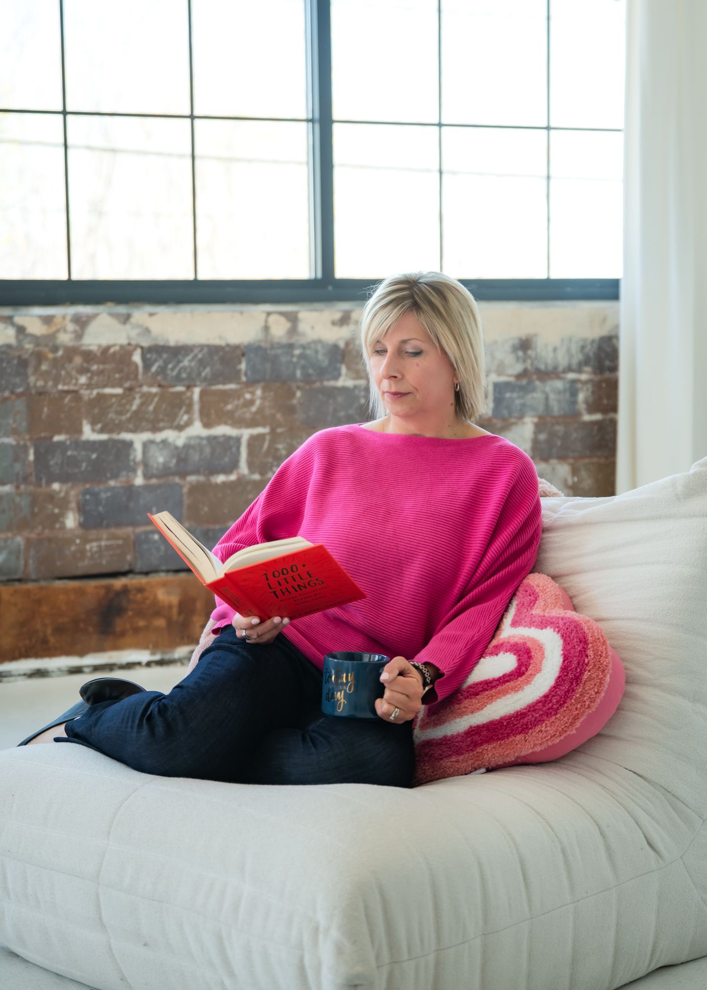 Woman in pink sweater reading a red book on a white couch with a mug, beside a brick wall and window.