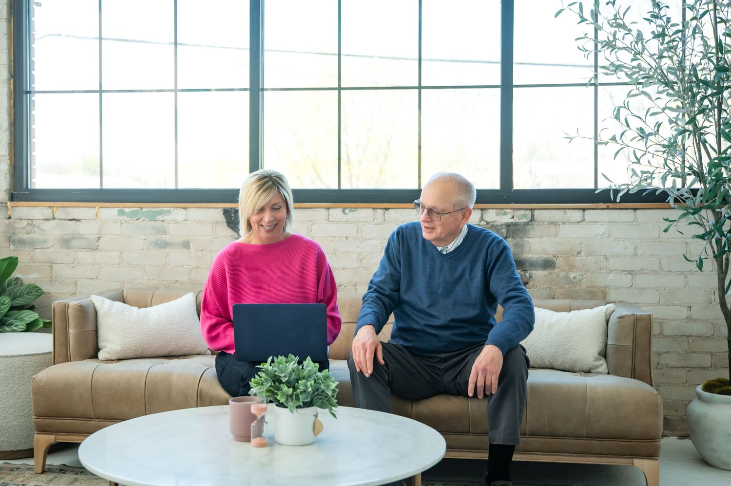 An older couple sits on a tan sofa, smiling at a laptop in a bright, modern living room with brick walls.