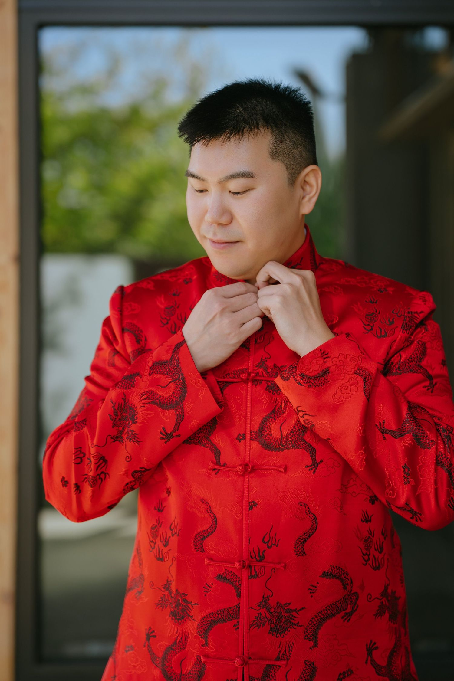 Groom in a red traditional Chinese jacket with embroidered dragons fastens his collar while getting ready for a wedding.