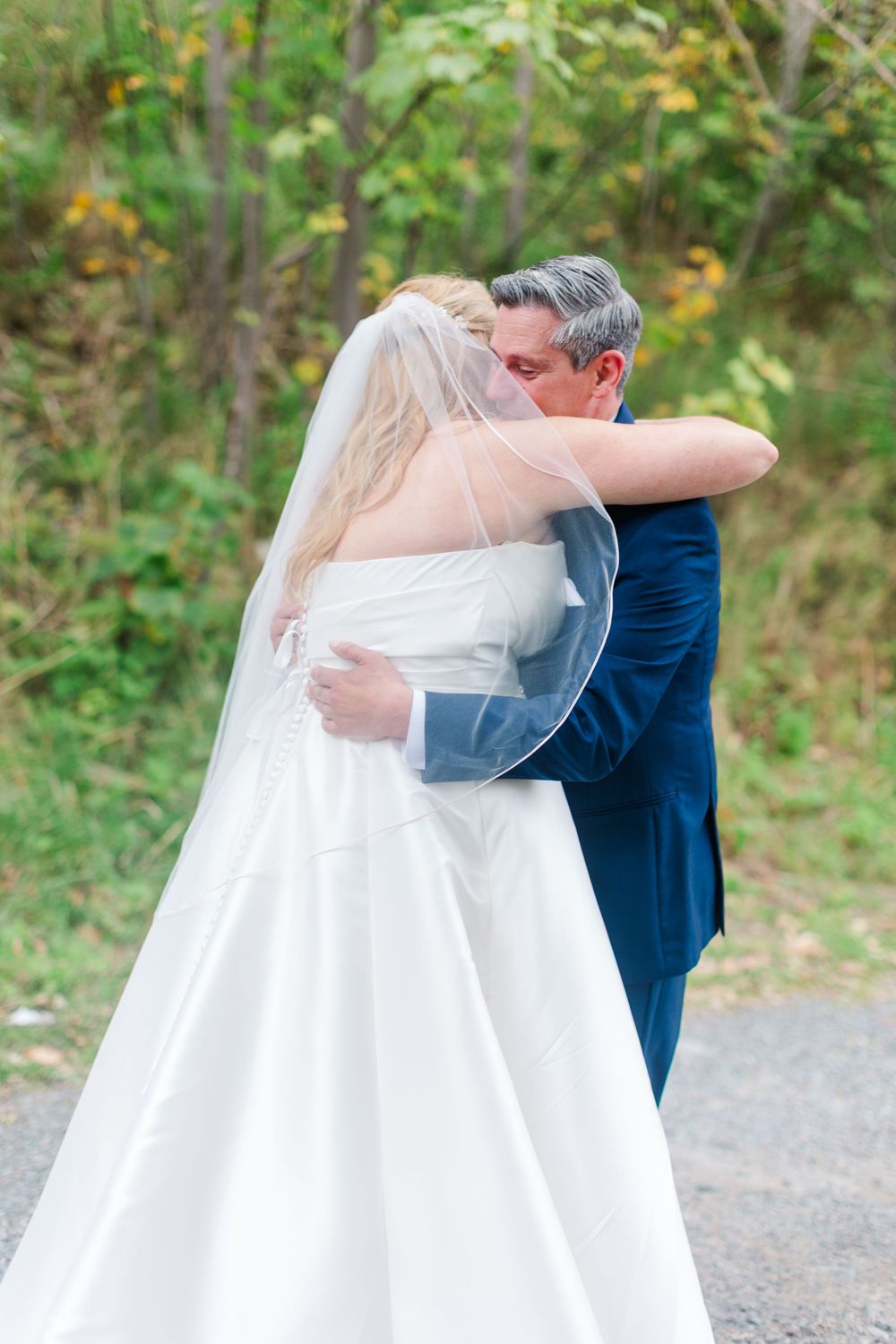 Bride in a white satin wedding gown and veil embraces a man in a navy suit on a gravel path with green and yellow foliage behind.