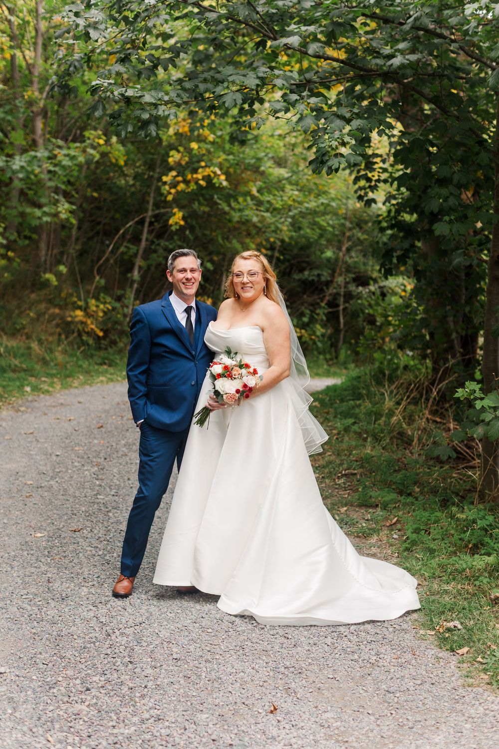 Smiling bride in a white strapless gown holding a bouquet stands beside a groom in a blue suit on a gravel path in a sunlit wooded setting.