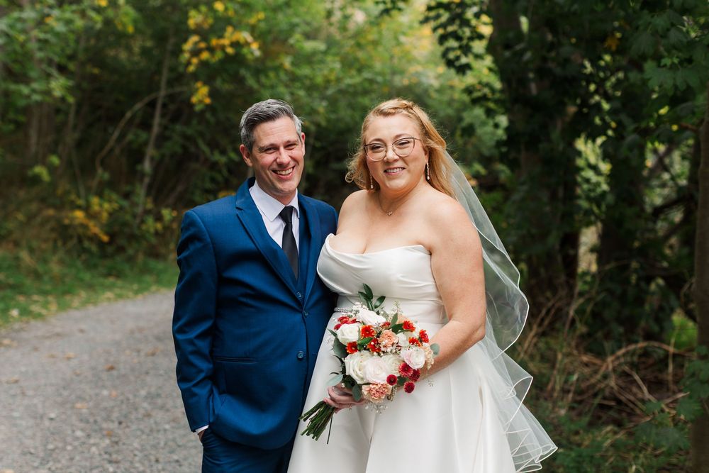 Smiling bride in a white strapless gown holding a red and white bouquet stands beside a groom in a blue suit on a wooded path.