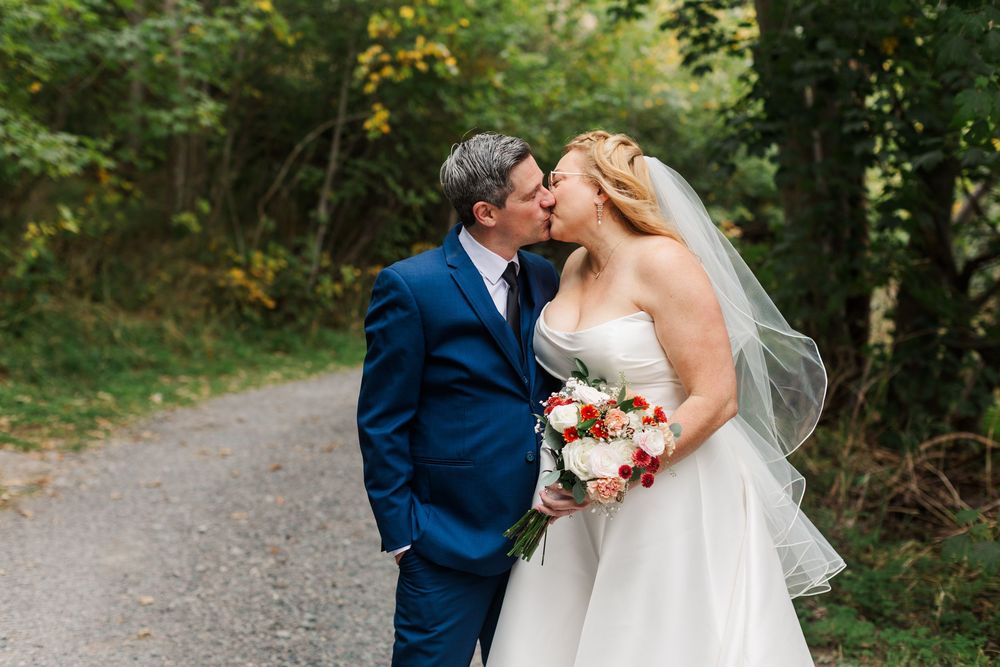 Bride in a white strapless gown and veil holding a bouquet kisses her groom in a navy suit on a gravel path surrounded by green trees.
