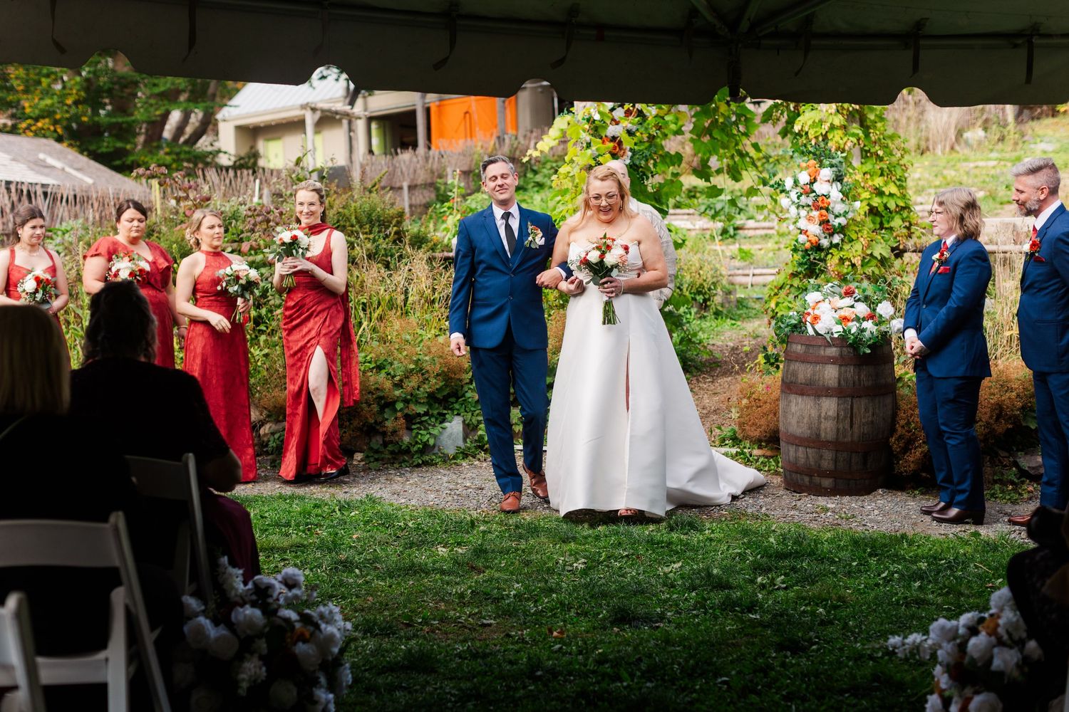 Bride in white gown and groom in navy suit walk hand in hand down the aisle at an outdoor garden wedding with bridesmaids in red dresses.