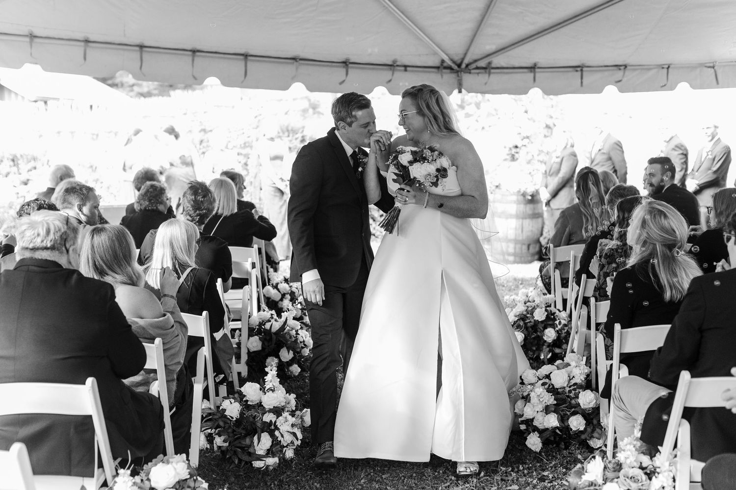 Bride and groom walk down the aisle together under a tent in a black and white outdoor wedding ceremony with guests and floral arrangements.