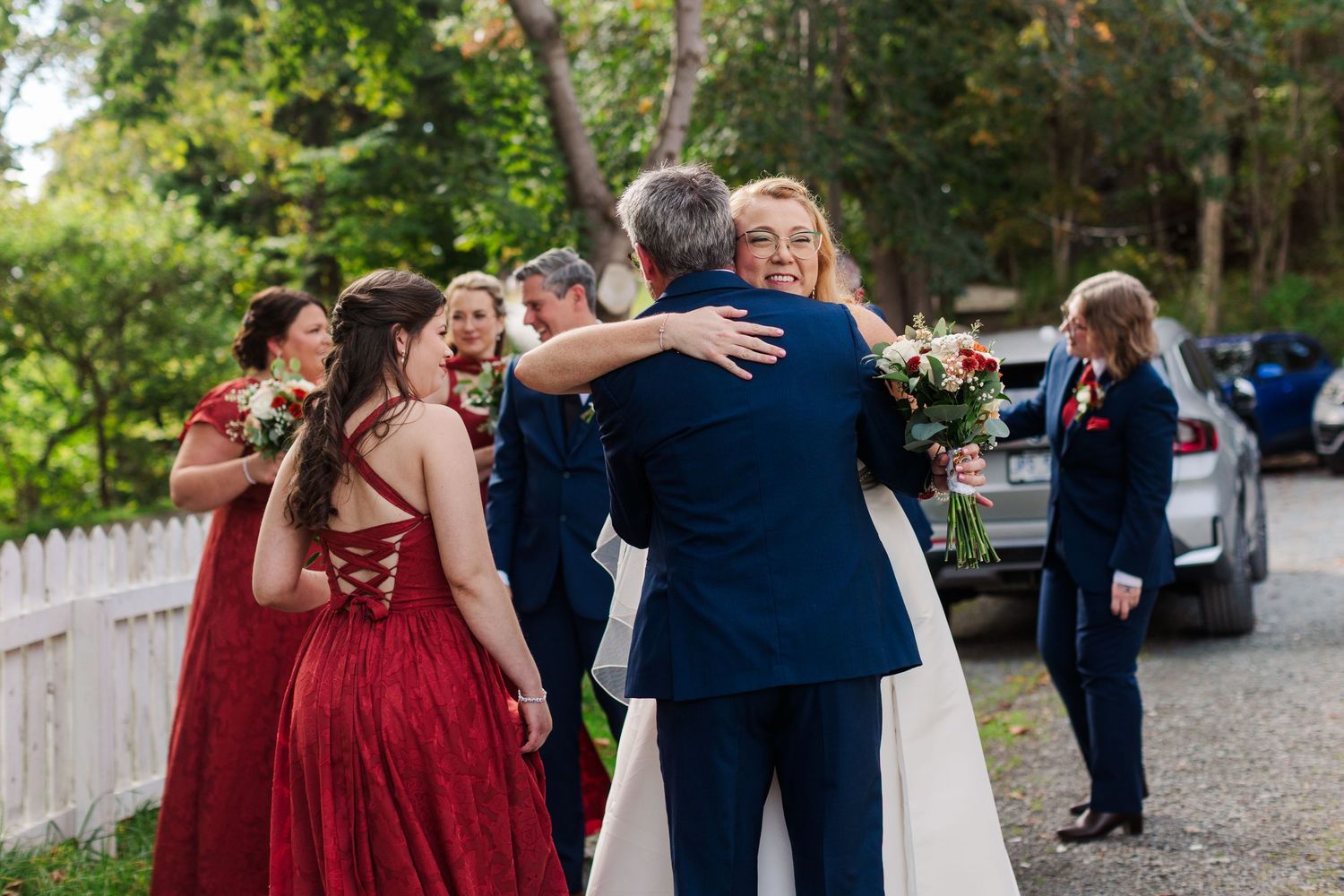 Bride in a white gown embraces a man in a navy suit while bridesmaids in red dresses hug nearby at an outdoor wedding.