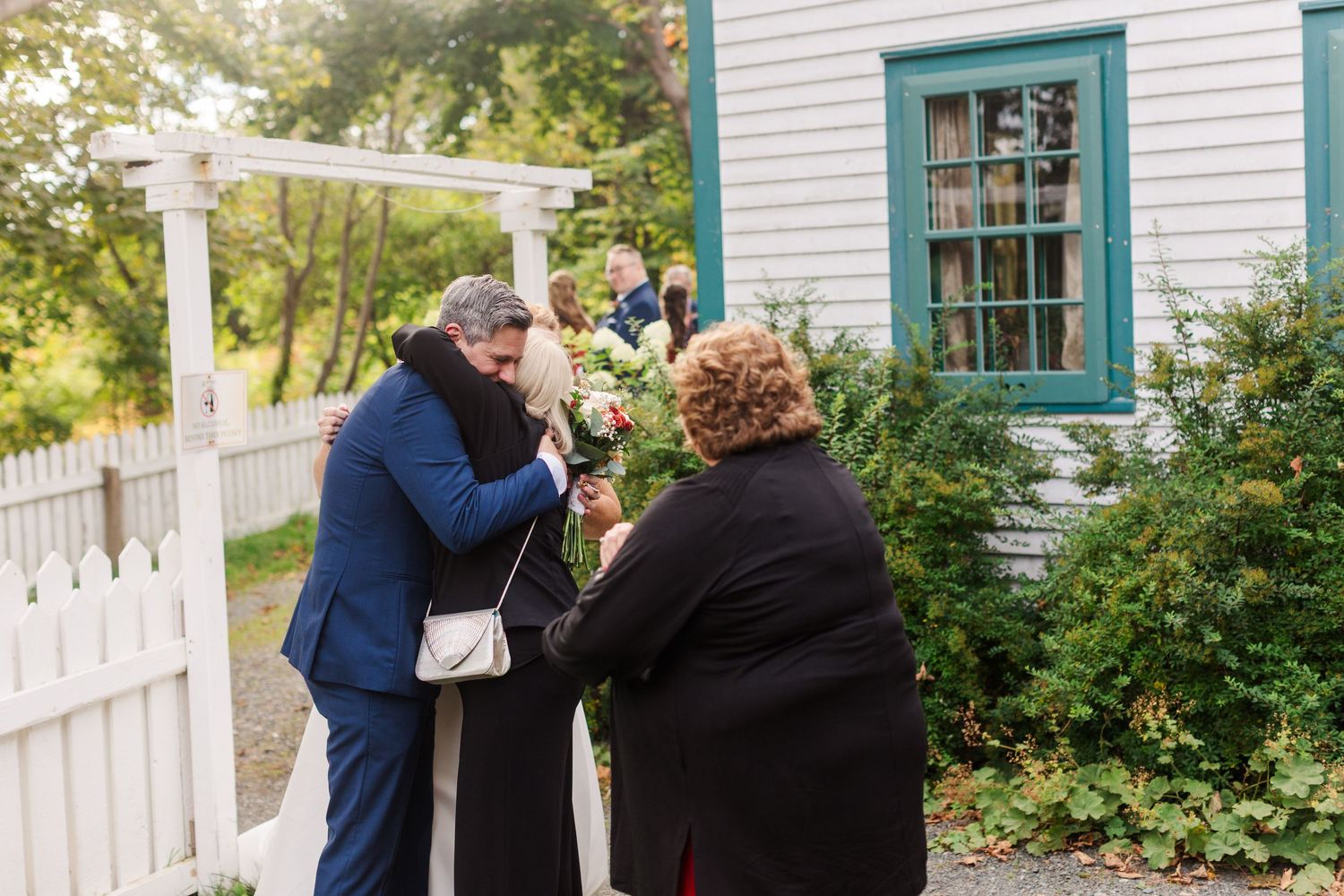 Wedding guests embrace and congratulate a couple near a white picket fence and gate outside a charming white cottage with green trim.