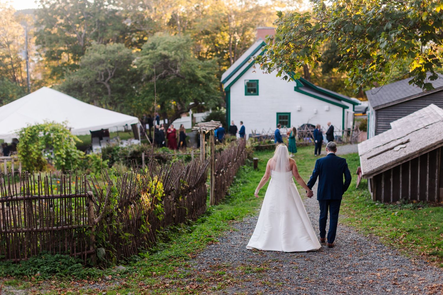 Bride in a white gown and groom in a dark suit hold hands walking down a gravel path toward guests at a rustic outdoor farm wedding.