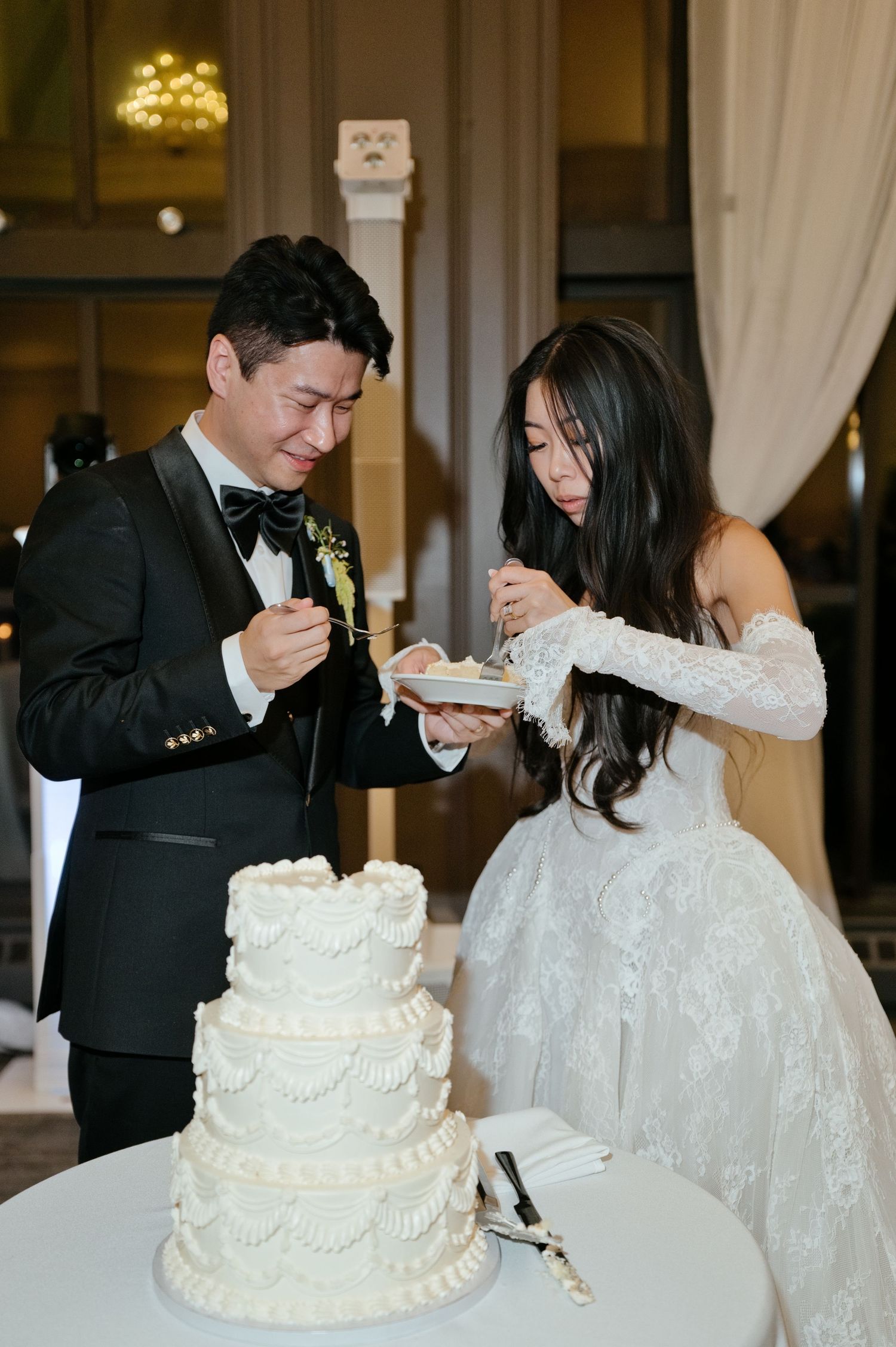 Bride and groom smiling as they share a slice of their white tiered wedding cake at the reception.