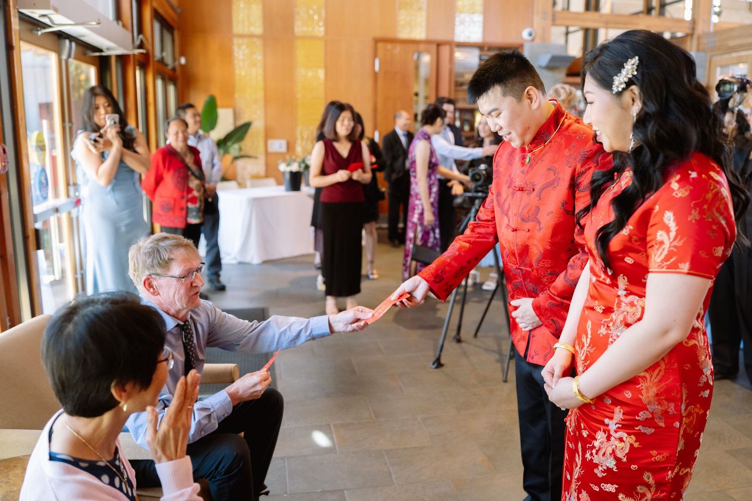 A couple in red traditional wedding attire receives a red envelope from a seated elder during a tea ceremony, while guests watch.