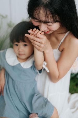 A smiling Asian mother in white dress gently kisses her toddler daughter's hands while the child looks forward.