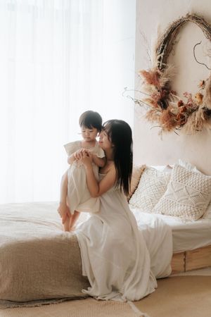 A mother in a white dress sits on a bed holding her young child, with a boho dried floral wreath on the wall.
