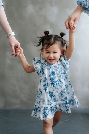 A joyful toddler girl in a blue floral dress holds hands with two adults, smiling brightly while learning to walk.