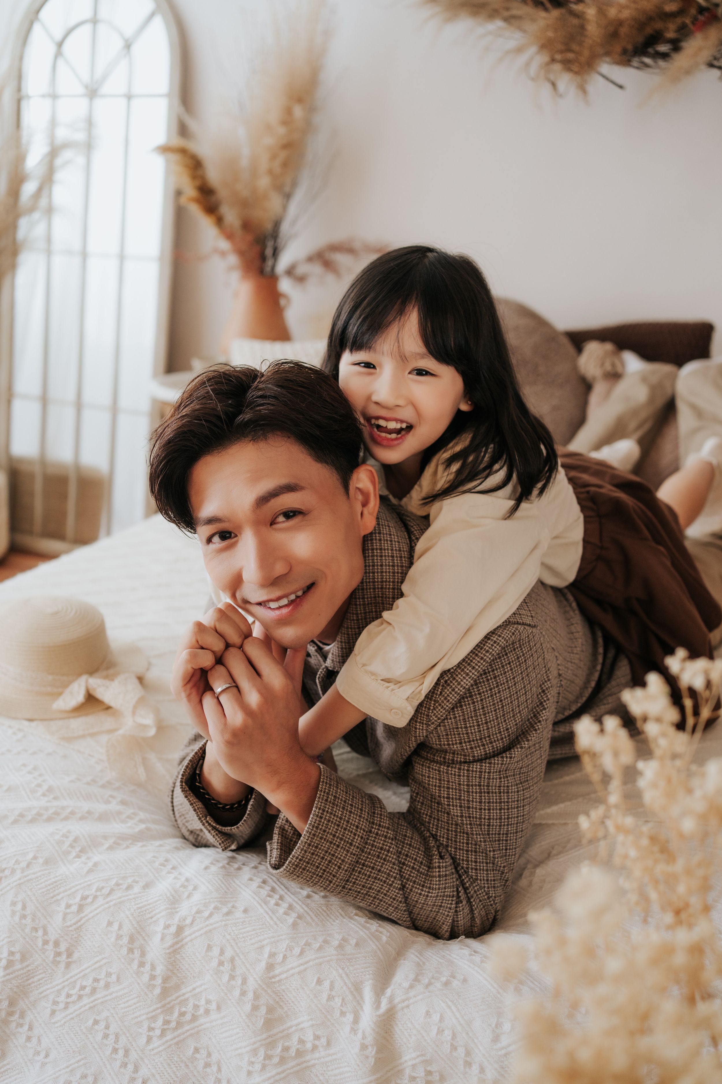 A smiling man and young girl laugh together while lying on a white bed in a cozy, warmly decorated room.
