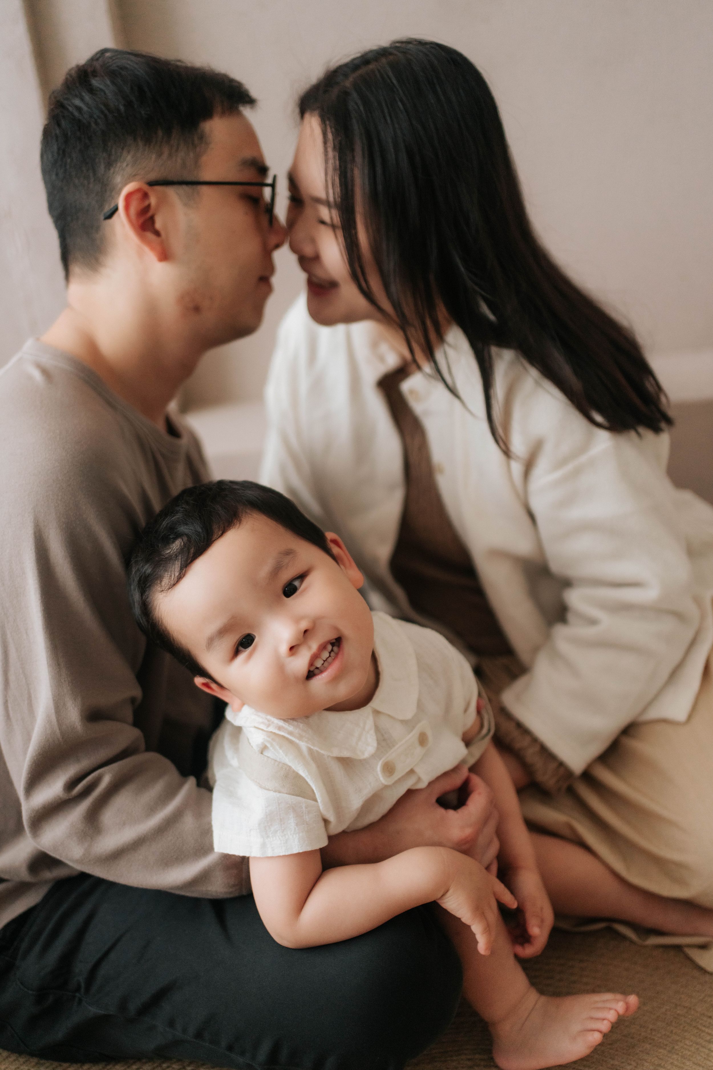 Asian family portrait with parents sharing a tender moment while their smiling baby sits between them.